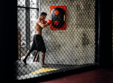 Sportsman with a naked torso and in the red boxing gloves hits equipment for boxing on a concrete wall in the gym behind the net .