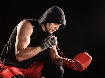 The young male athlete kickboxing sitting and lacing glove on a black  background