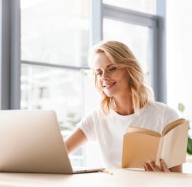 Smiling businesswoman working on laptop computer and holding open book while sitting at the office desk